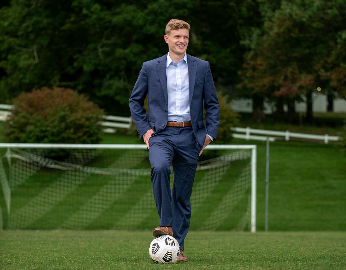 MariusGrytdal_soccerField student poses on soccer field in suit with foot on soccer ball